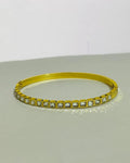 A gold-plated bangle bracelet featuring a row of square-cut clear stones, displayed on a neutral gray surface against a white background.