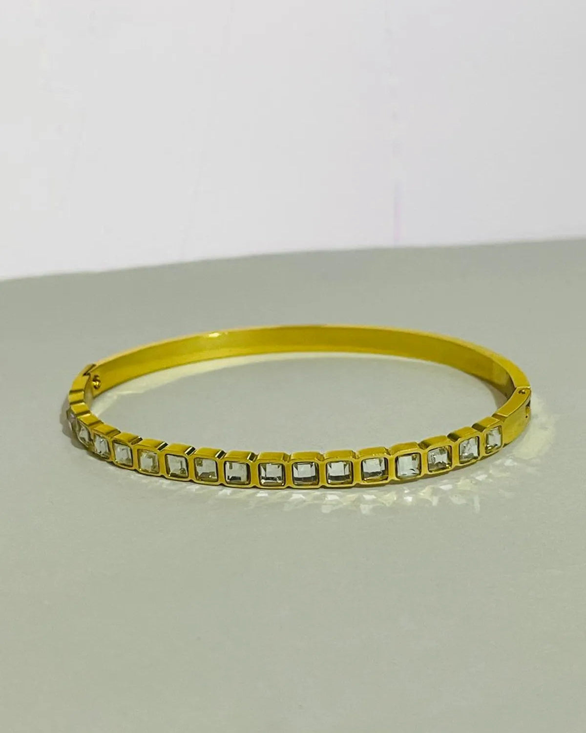 A gold-plated bangle bracelet featuring a row of square-cut clear stones, displayed on a neutral gray surface against a white background.