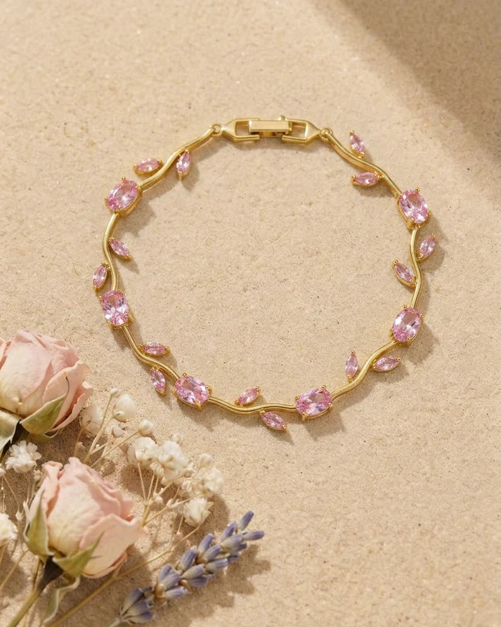 A gold chain bracelet adorned with small pink gemstones, arranged in a circle on a textured beige surface with dried pink roses and lavender flowers in the corner.